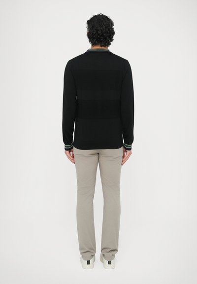 Čierny sveter s dlhými rukávmi s horizontálnymi pruhmi, zelenými a modrými akcentmi na golieri a manžetách, v kombinácii so svetlobéžovými nohavicami a bielymi topánkami.