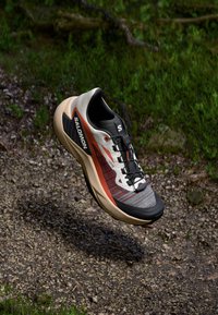 Salomon running shoe features a grey and orange mesh upper, black accents, a padded collar, and a lightweight beige outsole with grip.