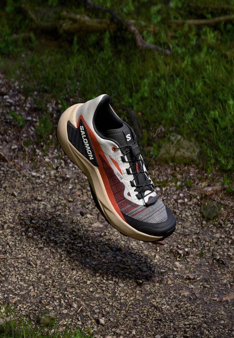 Salomon running shoe features a grey and orange mesh upper, black accents, a padded collar, and a lightweight beige outsole with grip.