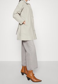 Veste de pluie légère beige à coupe droite, avec poches latérales et fermeture à boutons, associée à un jean gris à jambes larges et des bottines marron.