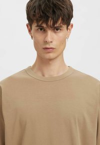 Pruun puuvillane t-särk, millel on ümmargune kaelus ja lühikesed varrukad, sileda tekstuuri ja lõdva lõikega. Lisandiks väike kuldne kõrvarõngas.