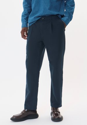 Chino blu navy con una vestibilità dritta, realizzati in tessuto misto cotone. Presentano un chiusura con bottone e pince frontali, abbinati a mocassini marrone scuro.