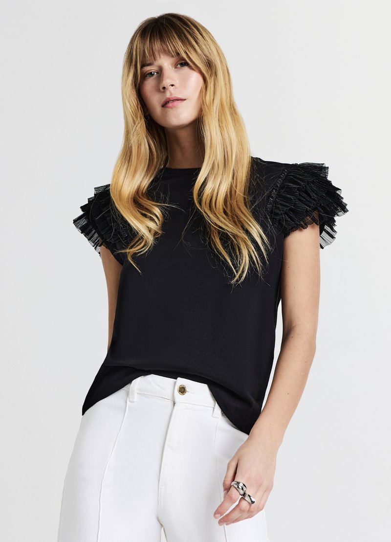 Haut noir à manches courtes avec des accents transparents et volantés sur les épaules ; associé à un pantalon blanc, présentant une coupe décontractée et une texture douce.