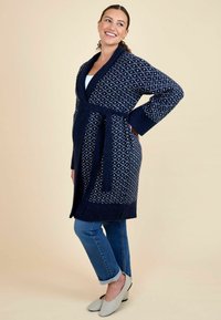 Cardigan tricotat de culoare bleu marin cu modele în diamant alb, cu centură în talie, cu guler tip șal și mâneci lungi, asortat cu blugi deschiși.