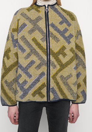 Personne portant une veste zippée à motifs jaunes, gris et noirs, de coupe oversize, avec un pantalon noir, debout devant un fond neutre.