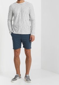 Chemise à manches longues gris clair avec de fines rayures horizontales, associée à un short bleu marine. Le modèle porte des baskets grises avec des accents blancs.