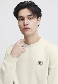 Hellbeige Sweatshirt mit Rundhalsausschnitt und gerippten Bündchen, versehen mit einem kleinen schwarzen Logopatch auf der linken Brust. Glatte Stoffstruktur.