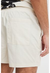 Beige shorts made of lightweight fabric, featuring an elastic waistband, side pockets, and a square back pocket, with a smooth texture.