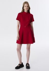 Abito rosso a maniche corte con collo alto e vita cinturata, caratterizzato da pieghe nella gonna. Indossato con sneakers nere legate.