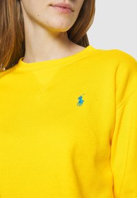 Gul sweatshirt med rund halsringning och ribbade ärmslut, med en liten blå logotyp på vänster bröst. Slät textur och avslappnad passform.