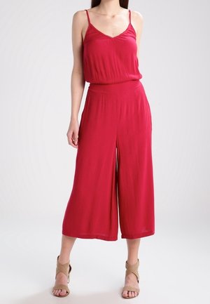 Kobieta ubrana w czerwony kombinezon bez rękawów z szerokimi, krótkimi spodniami oraz beżowymi sandałami na obcasie, stojąca na jednolitym tle.