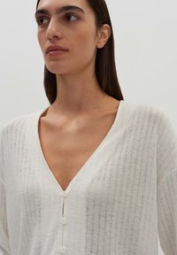 White knitted cardigan with a deep V-neck, featuring vertical rib patterns and small buttons along the front. Soft, lightweight texture.