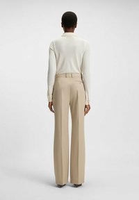 Beige tailored trousers with a straight leg, featuring a belt loop, delicate back zipper pocket, and a smooth finish. White long-sleeve top.