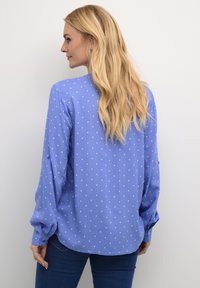 Blouse bleu clair à pois blancs, à manches longues, avec des poignets boutonnés, un léger éclat et un ourlet arrondi, associée à un jean bleu foncé.