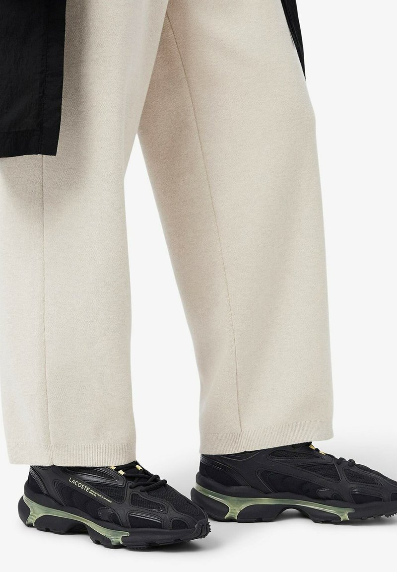 Chaussures de sport noires avec une tige texturée, dotées d'une semelle translucide verte. Associées à un pantalon ample beige clair.