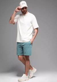 T-shirt blanc à manches courtes, shorts teal avec poches, et baskets beiges. Une casquette blanche avec un logo complète la tenue.