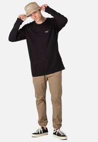 Hombre que lleva una camiseta de manga larga negra, pantalones beiges, zapatillas negras y un sombrero de cubo beige, ajustándose el sombrero en la cabeza.