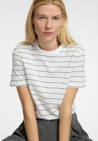 Vit kortärmad t-shirt med svarta horisontella ränder, rund halsringning, mjuk textur, bärs med grå byxor, minimalistisk design.
