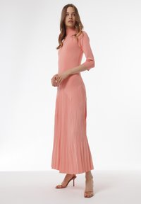 Lang, aansluitend jurkje in zacht koraal met een hoge halslijn en geribbelde textuur. Beschikt over een geplooide rok en zijopeningen voor bewegingsvrijheid.