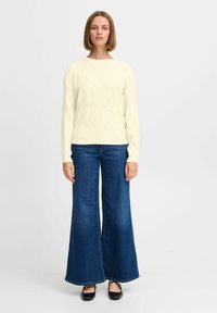 Světle béžový pletený svetr s kulatým výstřihem, spárovaný s širokými tmavými džíny. Měkká textura a jemné detaily na svetru.