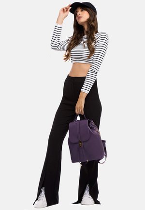 Zaino viola con pattina, hardware dorato e tasca frontale. Il modello indossa un top corto a righe bianche e nere e pantaloni neri con spacchi.