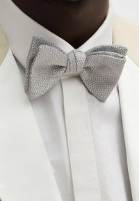 Primo piano di una persona che indossa un papillon argento texture sopra una camicia bianca e una giacca color crema.