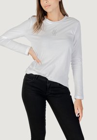 T-shirt bianco a maniche lunghe con scollo rotondo e un piccolo logo sul petto, abbinato a jeans neri slim-fit.