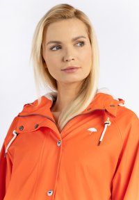 Narancssárga vízálló dzseki fehér gyapjúbéléses kapucnival, ezüst cipzárral és patentos gombokkal. Az elején egy ernyő logó található.
