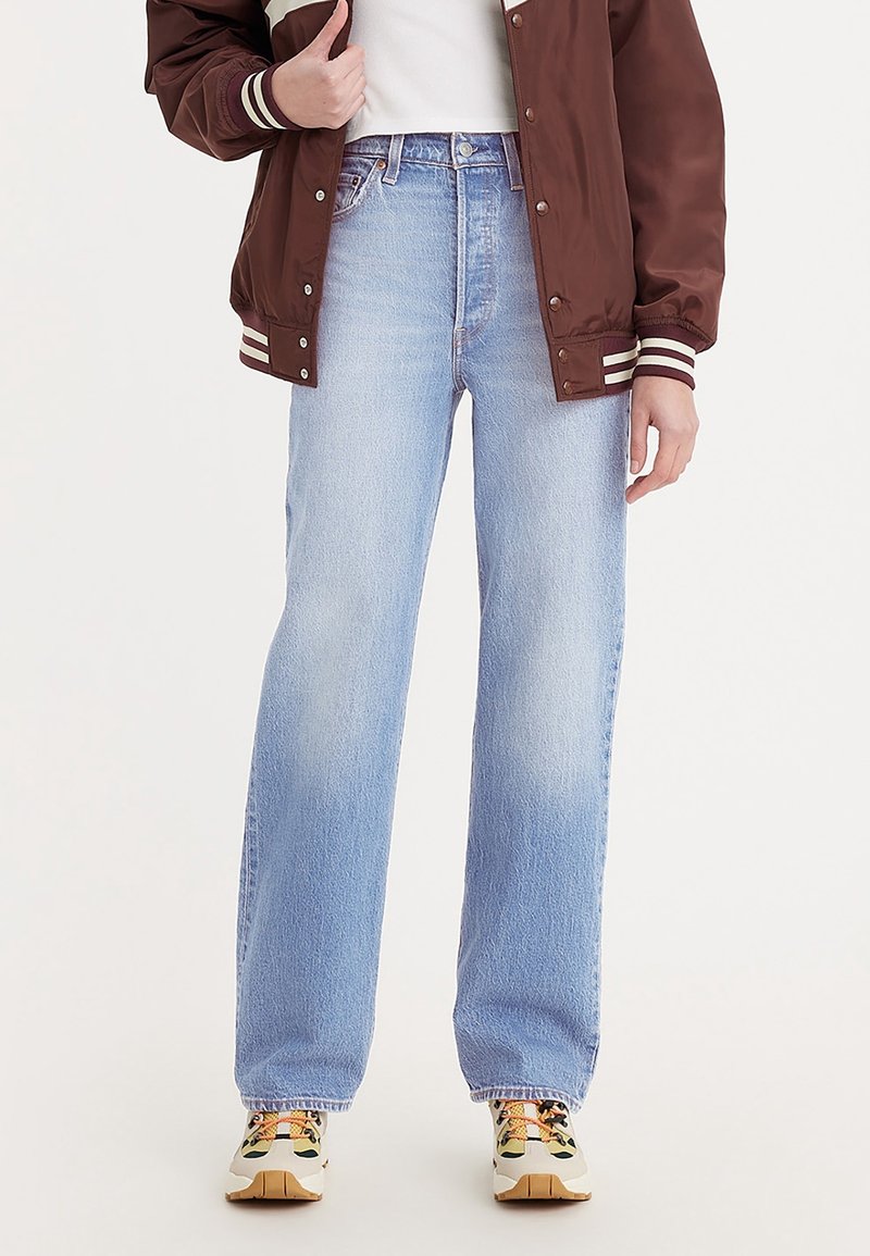 Personne portant un jean droit bleu clair, des baskets beiges et noires, une chemise blanche et une veste bomber marron avec des rayures blanches.