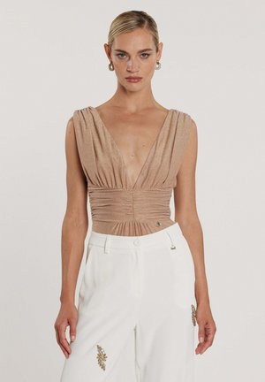 Donna che indossa un top beige senza maniche con scollatura profonda a V e pantaloni bianchi a vita alta con decorazioni dorate, in piedi davanti a uno sfondo semplice.