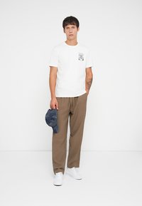 Maglietta bianca a maniche corte con grafica sul petto, pantaloni larghi in un tessuto marrone, e un berretto blu con motivo, abbinati a sneakers bianche.