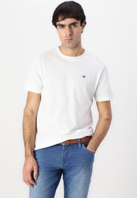 Camiseta de algodón blanca con cuello redondo y mangas cortas, con un pequeño logo en el pecho. Combinada con jeans de mezclilla azules y un cinturón marrón.