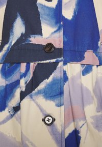 Kangas dengan pola sapuan kuas abstrak berwarna biru, ungu, dan merah jambu, dilengkapi dengan kancing biru navy dan putih pada bagian pleats pakaian.