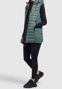 Kobieta w bezrękawniku w kolorze zielonym z kapturem, czarnej bluzce z długim rękawem, czarnych legginsach i czarnych sneakersach stoi na szarym tle.