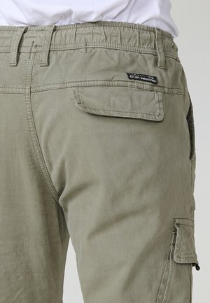 Šviesiai alyvuogių žalios spalvos cargo kelnės su elastinga juosta, su kišenėmis ant nugaros ir šonų, dėvimos su balta marškiniais.