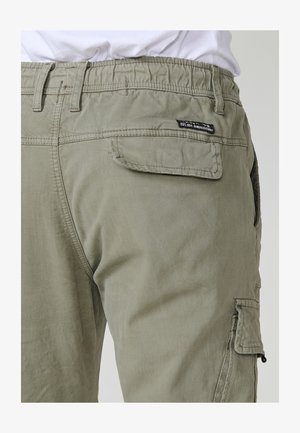 Világos olajzöld cargo nadrág elasztikus derékkal, hátul és oldalt hajtókás zsebekkel, fehér pólóval viselve.