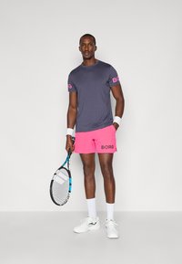 Sporta apģērbs, kurā ietilpst tumši zils īsai piedurknei krekls ar rozā "BORG" zīmolu, rozā šorti ar melnu "BORG" un baltas kurpes, kurās tur rokās tenisa raketi.