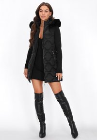 Czarna puchowa kamizelka o falistym wzorze, z kapturem z futerka, zamykanymi kieszeniami, noszona na czarnej sukience i wysokich czarnych botkach.