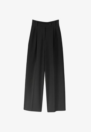 Femme debout de dos portant un haut blanc à manches courtes, un pantalon noir taille haute à jambes larges et des talons hauts beiges sur un fond uni.
