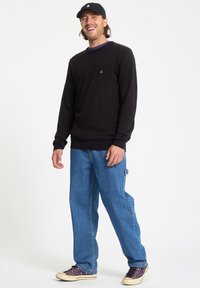Homme souriant, portant un t-shirt noir à manches longues, une casquette noire, un jean bleu et des baskets violettes, debout devant un fond blanc.