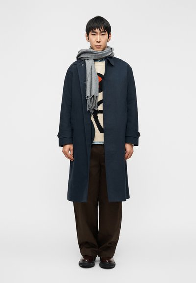 Homme debout portant un long manteau bleu marine, un pull à motifs, un pantalon large marron, des chaussures foncées et une écharpe grise enroulée autour de son cou.