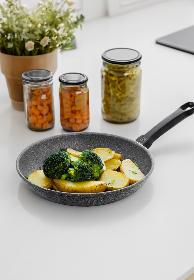 Poêle avec brocoli cuit et tranches de pommes de terre sur un plan de travail blanc, à proximité des bocaux de carottes et de cornichons, ainsi qu'une petite plante en pot.