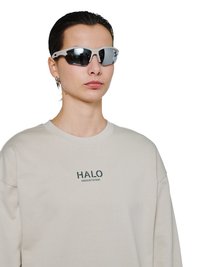 Person iført hvide sports-solbriller og beige sweatshirt med teksten "HALO", vist fra brystet og op mod en hvid baggrund.
