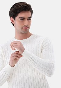 Weißer Kabelstrickpullover mit gerippten Bündchen und Halsausschnitt. Hat strukturierte Muster aus einem weichen, leichten Stoff. Nahaufnahme von Händen.