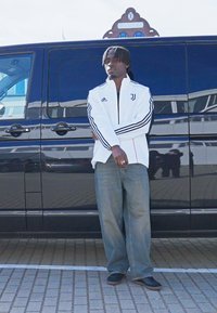 Sportsjakke i hvid med sorte striber, logo på brystet; løse blå jeans og sorte sko, stående ved siden af en sort varevogn.