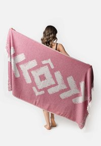 Gran manta rosa con patrones geométricos blancos y bordes con flecos. Textura suave y forma rectangular, sostenida horizontalmente por una persona.