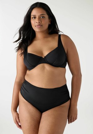 Donna con capelli lunghi e scuri che indossa un top bikini nero con ferretto e slip bikini a vita alta, in piedi contro uno sfondo chiaro e uniforme.