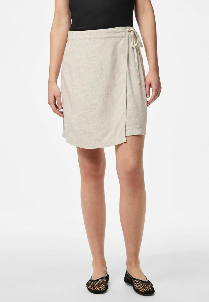 Spódnica o długości do kolan, stylu kopertowym, w jasnym beżowym lnie, z wiązanym pasem i subtelną teksturą. Noszona z czarnymi balerinami z siateczki.