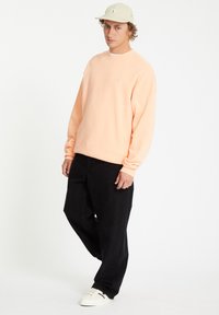 Sweatshirt de couleur pêche avec col rond, associé à un pantalon noir ample et des baskets blanches. La casquette est de couleur beige clair avec un logo.