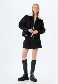 Schwarze, cropped Jacke mit cremefarbenem Lammfellkragen, kombiniert mit einem schwarzen, plissierten Rock und knielangen, schwarzen Lederstiefeln mit klobigen Sohlen.